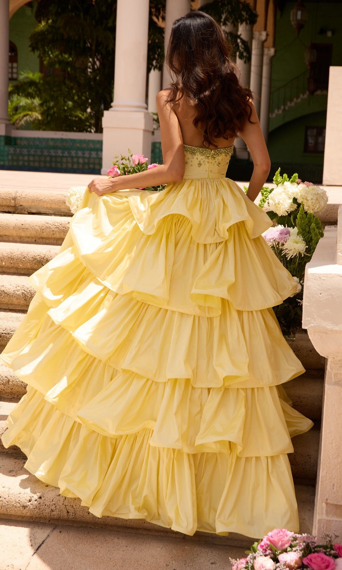 A woman wearing the Ava Presley 26417 Long Prom Dress, a strapless tiered yellow ruffle ball gown, stands on outdoor steps holding her dress, surrounded by flowers and greenery.